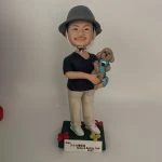 一款站立的人像公仔置於深綠色方形底座上，戴灰色漁夫帽、黑色上衣、米色長褲與白色球鞋；雙手懷抱一隻穿藍綠背帶的棕色捲毛小型犬寵物公仔，犬隻五官圓潤、耳朵下垂，呈現貴賓犬風格。底座四角有紅黃小花裝飾，前方附白色名牌刻字。背景為素白。