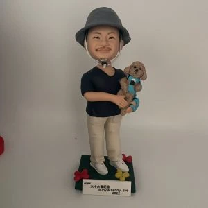 一款站立的人像公仔置於深綠色方形底座上，戴灰色漁夫帽、黑色上衣、米色長褲與白色球鞋；雙手懷抱一隻穿藍綠背帶的棕色捲毛小型犬寵物公仔，犬隻五官圓潤、耳朵下垂，呈現貴賓犬風格。底座四角有紅黃小花裝飾，前方附白色名牌刻字。背景為素白。