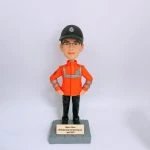 救護主題的人像公仔站在灰色石紋方形底座上，雙手叉腰；公仔戴黑色帽、配眼鏡，穿亮橘色高可視外套，衣身與袖口有銀色反光條與臂章徽章，下著黑色長褲與黑鞋。底座前有金色小牌，刻有英文姓名與「Ambulancing man passing out April 2023」字樣。