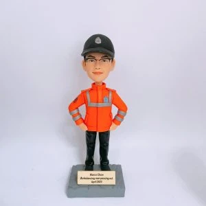 救護主題的人像公仔站在灰色石紋方形底座上，雙手叉腰；公仔戴黑色帽、配眼鏡，穿亮橘色高可視外套，衣身與袖口有銀色反光條與臂章徽章，下著黑色長褲與黑鞋。底座前有金色小牌，刻有英文姓名與「Ambulancing man passing out April 2023」字樣。