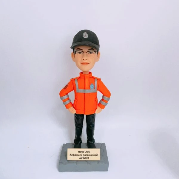 救護主題的人像公仔站在灰色石紋方形底座上，雙手叉腰；公仔戴黑色帽、配眼鏡，穿亮橘色高可視外套，衣身與袖口有銀色反光條與臂章徽章，下著黑色長褲與黑鞋。底座前有金色小牌，刻有英文姓名與「Ambulancing man passing out April 2023」字樣。