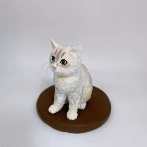 單隻白奶油色相間的坐姿貓公仔置於圓形棕色底座上，粉色鼻頭、黑大眼、耳尖立起；毛流以細緻刻線呈現，鬍鬚纖細可見。背景為簡潔淺灰白色。