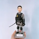 一款穿著灰色戰術服的人像公仔站在灰色方形底座上：身著長袖連身服，外配黑色戰術背心與多個裝備袋，胸前有黃色扣件，腰帶與大腿處掛載裝備；雙膝配護膝、足穿黑色鞋，右手握有細長裝備棒狀物。左上臂見紅色臂章，整體呈現消防或特勤救援風格。底座前方貼有白色名牌。背景為淺藍色牆面，前景可見手掌托著底座。