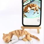 一隻橘白虎斑家貓造型的寵物公仔側躺熟睡在白色背景上，橘色虎斑與白胸、白前腳、白後腳細節清晰，尾巴自然向後延伸。後方有一支立在小木架上的手機，螢幕顯示原貓照片：同樣的橘白虎斑家貓以相同姿勢睡在金屬桌面上，呈現照片到公仔的對照。