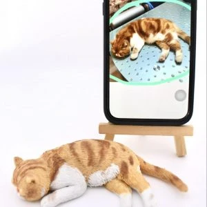 一隻橘白虎斑家貓造型的寵物公仔側躺熟睡在白色背景上，橘色虎斑與白胸、白前腳、白後腳細節清晰，尾巴自然向後延伸。後方有一支立在小木架上的手機，螢幕顯示原貓照片：同樣的橘白虎斑家貓以相同姿勢睡在金屬桌面上，呈現照片到公仔的對照。