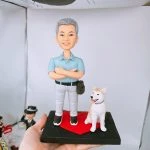 手持黑色底座的人像公仔擺設：灰白短髮男子公仔雙臂交叉，著淺藍短袖襯衫、淺灰長褲與黑色運動鞋，側掛咖啡色小包；身旁坐著一隻白底淡棕斑點的中型犬公仔，吐舌微笑。兩者站在紅色愛心形平台上，背景為簡潔白色棚拍環境。
