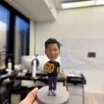 採用站立姿勢，雙手持有一個醒目的金色「60」數字，象徵著重要的里程碑與慶祝時刻。 公仔穿著 黑色休閒上衣 搭配 深色長褲，腳踩 運動鞋，展現簡約隨性的日常風格；髮型整潔俐落，神情帶有微笑，塑造出親切又穩重的形象。 底座以 簡潔圓形灰色設計，讓整體焦點更集中於公仔與「60」的慶祝元素。