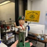 童軍主題人像公仔站在綠色方形底座上，戴綠色貝雷帽與徽章，穿卡其色短袖制服配紅色領巾、綠色長褲與黑鞋，右手高舉、左手持白色旗桿，旗面為黃色並印有童軍百合花徽與英文隊名。