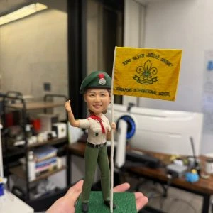 童軍主題人像公仔站在綠色方形底座上，戴綠色貝雷帽與徽章，穿卡其色短袖制服配紅色領巾、綠色長褲與黑鞋，右手高舉、左手持白色旗桿，旗面為黃色並印有童軍百合花徽與英文隊名。