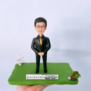 榮休主題的人像公仔：戴眼鏡的短髮男士身穿黑色西裝與金黃色領帶，雙手於身前交握，站在綠色草地質感的地台上；地台前緣設有兩塊金屬銘牌，其一寫有「10年最佳拍檔」，另一塊標示「Mr. Donald Fan Happy Retirement 2022」與祝福字樣。左後方有白色指示牌小道具，右前方擺放以編織圈與綠植構成的小擺飾；整體以淺灰白背景拍攝，手掌托起作品，呈現適合作為高層榮休禮物的人像公仔展示。