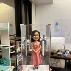 一款人像公仔置於透明壓克力盒內：女性公仔留中長黑髮、戴黑框眼鏡，穿粉色無袖長洋裝與深色高跟鞋，頸上配長項鍊，雙手於身前交握，站在白色圓形底座上。底座前有金色名牌，刻字含「Mrs Ng」「We love our English teacher」「2023」「3D students, Ms Yam & Mr Leung」等英文字樣。背景為辦公室/教職員室環境，可見桌面、文件與架子等雜物。