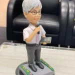 宗教主題的人像公仔：銀灰短髮、戴眼鏡的男性站姿，穿白襯衫配淡紫色領帶與深灰長褲，右手握麥克風，左手持黑色金邊聖經樣小冊；腳踏石磚紋台座，四周綠邊點綴小花。適合作為牧師禮物的人像公仔。