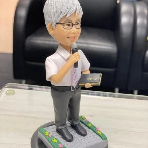 宗教主題的人像公仔：銀灰短髮、戴眼鏡的男性站姿，穿白襯衫配淡紫色領帶與深灰長褲，右手握麥克風，左手持黑色金邊聖經樣小冊；腳踏石磚紋台座，四周綠邊點綴小花。適合作為牧師禮物的人像公仔。