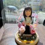 教師主題人像公仔置於木質圓形底座中央，底座內圈繞有暖黃燈串；公仔穿米色外套與碎花襯衫、長髮微卷微笑站立。四周以粉紅與玫瑰色不凋花、苔蘚與小白花環繞，後方形成粉色花環拱門；前方金色小牌寫有「謝謝你無私的指導」。左側放著透明玻璃罩，背景是靠窗的城市景致與校園般建築。