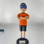 休閒風格的人像公仔雙臂交叉站立於黑色圓形底座上，戴反戴灰色帽、配大型反光墨鏡，穿橘色短袖上衣、深色及膝短褲、藍白襪與黑紅運動鞋；底座前貼有小牌寫著「Dear Wa Happy Birthday Sep 2021」。