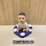 木紋背景前，一款小朋友主題bb人像公仔坐在紫色心形底座上：bb採盤腿坐姿，穿著彩色圖案連身衣，表情溫和；心形底座表面具細砂紋理並環繞多朵白色玫瑰造型小花。前方擺放白色長條名牌，上寫「Cutefigure_hk」。