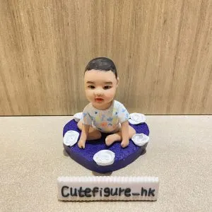 木紋背景前，一款小朋友主題bb人像公仔坐在紫色心形底座上：bb採盤腿坐姿，穿著彩色圖案連身衣，表情溫和；心形底座表面具細砂紋理並環繞多朵白色玫瑰造型小花。前方擺放白色長條名牌，上寫「Cutefigure_hk」。