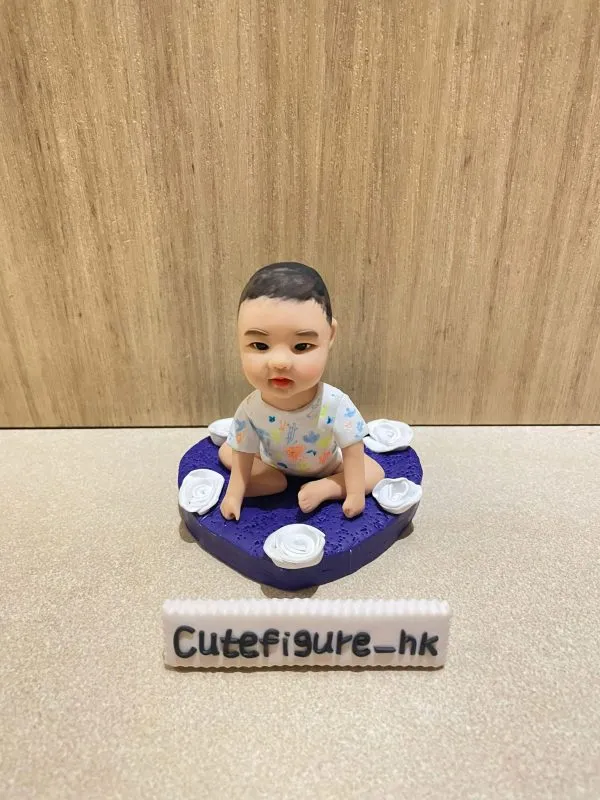 木紋背景前，一款小朋友主題bb人像公仔坐在紫色心形底座上：bb採盤腿坐姿，穿著彩色圖案連身衣，表情溫和；心形底座表面具細砂紋理並環繞多朵白色玫瑰造型小花。前方擺放白色長條名牌，上寫「Cutefigure_hk」。
