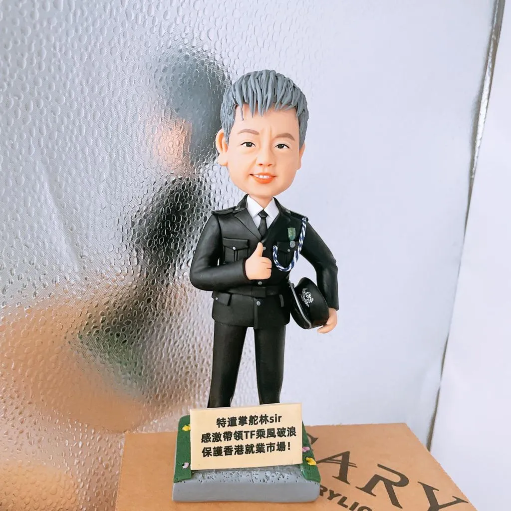 深色禮服風格的警察主題人像公仔站在灰色底座上，右手比讚、左手持有帶徽章的制帽；肩上垂掛藍白肩繩，外套口袋與徽章細節清晰。底座前方立有中文感謝字牌。背景為磨砂質感半透明板與紙盒邊角。
