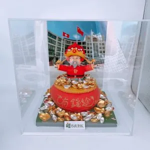 一座以財神造型為主題的人像公仔置於透明展示盒內：公仔穿紅袍戴金色大帽、留灰白鬍鬚，雙臂張開站在大紅聚寶盆上，周圍堆滿金色與銀色元寶。前方貼有「EdUHK」字樣的小牌；背景為一幅校園建築與多面旗幟的照片。