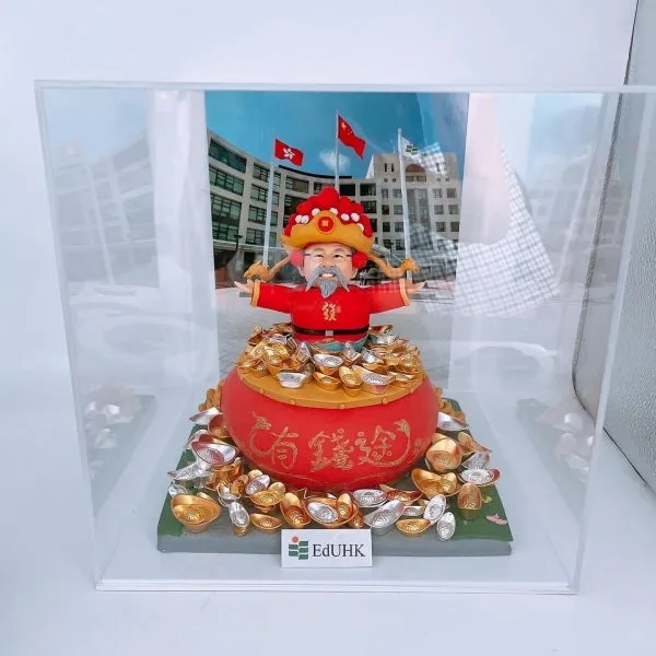 一座以財神造型為主題的人像公仔置於透明展示盒內：公仔穿紅袍戴金色大帽、留灰白鬍鬚，雙臂張開站在大紅聚寶盆上，周圍堆滿金色與銀色元寶。前方貼有「EdUHK」字樣的小牌；背景為一幅校園建築與多面旗幟的照片。