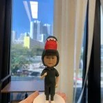 一款小朋友／bb主題的人像公仔立於白色圓形底座上，被手掌托著展示。公仔穿全黑連身服與黑色安全帽，帽頂裝飾紅色帶白齒狀邊的恐龍頭造型；腳穿白底粉邊鞋，胸前綁有黑色蝴蝶結。背景是大窗外的城市高樓與藍天，室內有米色窗簾與木桌。