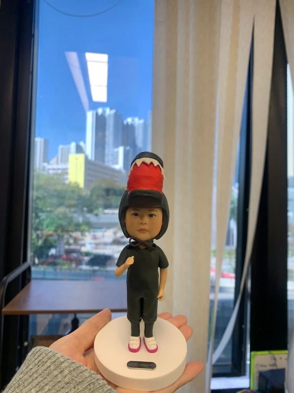 一款小朋友／bb主題的人像公仔立於白色圓形底座上，被手掌托著展示。公仔穿全黑連身服與黑色安全帽，帽頂裝飾紅色帶白齒狀邊的恐龍頭造型；腳穿白底粉邊鞋，胸前綁有黑色蝴蝶結。背景是大窗外的城市高樓與藍天，室內有米色窗簾與木桌。