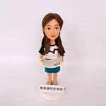 女老師主題的人像公仔站在白色圓形底座上，長髮披肩、面帶微笑，穿黑白短袖上衣與藍色及膝裙，雙手持展開的樂譜紙；底座前貼有小牌，寫著「感激遇到好老師」。純白背景拍攝。