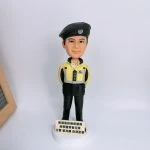 一款聖約翰救傷隊主題的人像公仔直立於白色圓形底座上：角色戴黑色貝雷帽，上有徽章圖案；身穿黃黑相間的制服外套與黑色長褲，雙手放於身後，胸前配有名章與口袋細節。底座前方立有小牌，印有「香港聖約翰救傷隊」及隊屬文字。背景為素白牆面，左側可見木框一角。