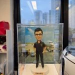 男性人像公仔置於透明展示盒內，站在白色圓形底座上；公仔戴黑框眼鏡，短髮，穿深色短袖工作POLO衫與長褲、黑鞋，胸前有盾形徽章與小國旗樣式貼章，一手插袋一手自然下垂。底座前方貼有小牌寫著「感謝你根哥 退休生活愉快」。