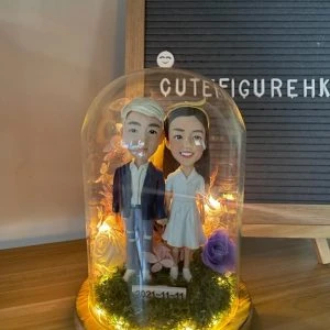 木質底座上的玻璃永生花燈罩作品：內含一對情侶人像公仔，左側角色著深藍外套與破洞牛仔褲、白鞋；右側角色穿白色V領短袖連身裙與白鞋。腳下覆苔綠草，周圍點綴紫玫瑰與乾燥花材，環繞暖白色小燈串；前方小牌寫有日期「2021-11-11」。背景為木桌與置有字母板的場景。