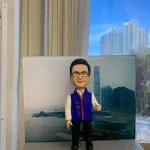 男性人像公仔站在灰色長方形底座上，背後為海港與高樓天際線背景板；公仔戴黑框眼鏡、短髮，穿白襯衫與紫色背心、黑色長褲與黑鞋，胸前掛著紅藍邊頸帶獎章。底座側貼有白色標牌，印有黑字「孔議員，辛苦你了！」；畫面右側可見手掌托住底座。