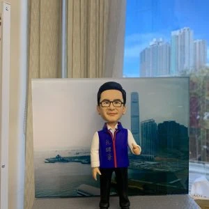 男性人像公仔站在灰色長方形底座上，背後為海港與高樓天際線背景板；公仔戴黑框眼鏡、短髮，穿白襯衫與紫色背心、黑色長褲與黑鞋，胸前掛著紅藍邊頸帶獎章。底座側貼有白色標牌，印有黑字「孔議員，辛苦你了！」；畫面右側可見手掌托住底座。