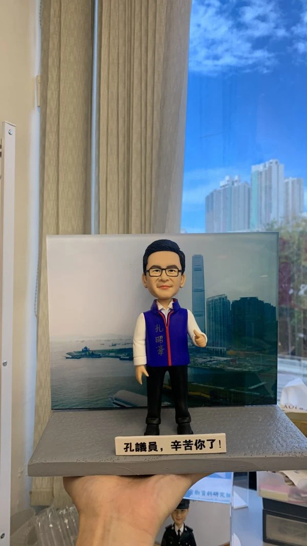 男性人像公仔站在灰色長方形底座上，背後為海港與高樓天際線背景板；公仔戴黑框眼鏡、短髮，穿白襯衫與紫色背心、黑色長褲與黑鞋，胸前掛著紅藍邊頸帶獎章。底座側貼有白色標牌，印有黑字「孔議員，辛苦你了！」；畫面右側可見手掌托住底座。