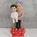 水泥灰背景前，一組祖孫主題人像公仔立於紅色心形底座：年長角色穿粉棕色針織上衣與黑褲、米色鞋，面帶微笑從後方雙手托起懷中的bb；小朋友穿白色點點襯衫與白色褲襪，短髮、神情可愛。底座撒有粉、黃小花瓣裝飾，前方紅色小牌寫「CuteFigureHK」。
