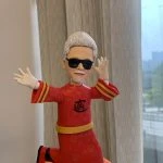 戴黑色墨鏡、白色短髮與小鬍子的男性人像公仔，單膝前跨、雙臂張開的動感姿勢；身穿紅色中式長袍，衣身帶有低飽和暗花紋理，胸前為黑色書法風圖案，袖口三道金色條紋，腰間繫金色腰帶，腳穿紅黑靴。公仔站在橘色多孔質感的圓形底座上；背景為室內窗簾與窗外街景，有人手掌托住底座。