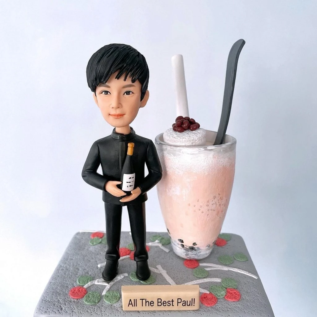 高檔餐廳侍應主題的人像公仔：短黑髮年輕男性穿著全黑侍應制服（立領上衣與長褲），雙手於腹前抱持一瓶酒；公仔站在灰色紋理底座上，表面點綴紅綠圓點與白色枝條圖樣。右側擺放一杯大型粉色奶昔／調飲模型，頂部有鮮奶油與紅豆造型，插著白色餅乾棒與黑色長匙。前方木質小牌刻有「All The Best Paul!」。背景為簡潔白牆，整體呈現高檔餐廳工作人員（侍應）致敬或餞別禮的人像公仔。