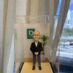 辦公室主題的人像公仔：一位外國男性老闆形象，光頭、微笑表情，穿黑色西裝外套與白襯衫、藍色反摺長褲與棕色短靴，雙手於腹前相合站姿。公仔置於木紋地台，後方有綠植與牆上綠白色公司標誌牌，整體收納在透明防塵展示盒中；背景為落地窗簾與城市景觀。作品呈現適合作為老闆或上司的祝福禮物的人像公仔。
