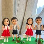 桌面上兩組小朋友主題人像公仔並排展示：每組各有一位女孩與一位男孩，皆以Q版比例站在草綠不規則底座上。女孩穿白襯衫搭紅色吊帶裙與白鞋，雙手比出V手勢；男孩穿白底黑格襯衫配黑色吊帶短褲與深色鞋，同樣比V手勢。每個底座上都點綴白色小指示牌、石頭與棕色編織小籃盛放綠色葉材；背景為室內窗邊與淺色窗簾，窗外高樓景色散景。