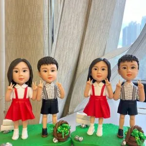 桌面上兩組小朋友主題人像公仔並排展示：每組各有一位女孩與一位男孩，皆以Q版比例站在草綠不規則底座上。女孩穿白襯衫搭紅色吊帶裙與白鞋，雙手比出V手勢；男孩穿白底黑格襯衫配黑色吊帶短褲與深色鞋，同樣比V手勢。每個底座上都點綴白色小指示牌、石頭與棕色編織小籃盛放綠色葉材；背景為室內窗邊與淺色窗簾，窗外高樓景色散景。