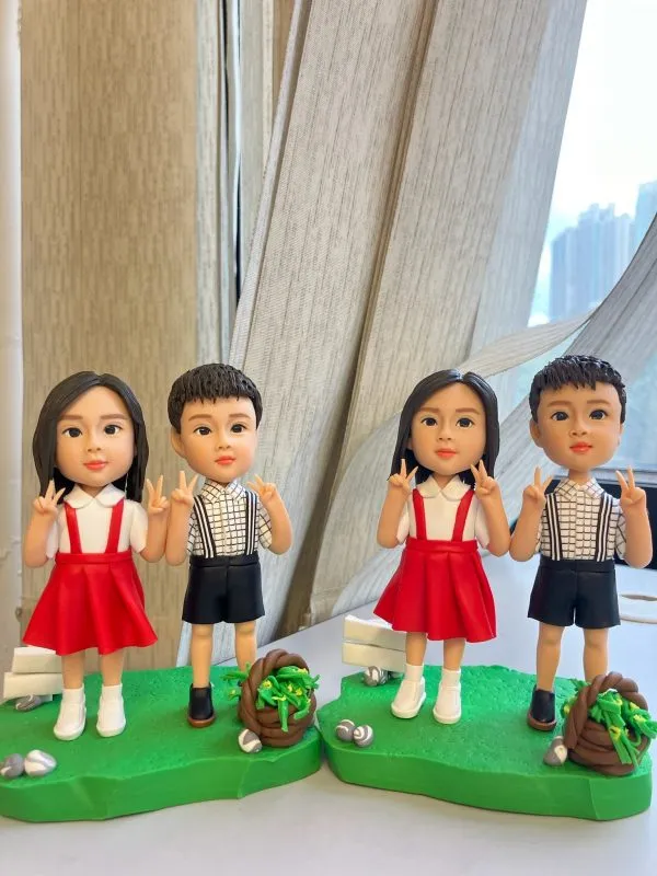 桌面上兩組小朋友主題人像公仔並排展示：每組各有一位女孩與一位男孩，皆以Q版比例站在草綠不規則底座上。女孩穿白襯衫搭紅色吊帶裙與白鞋，雙手比出V手勢；男孩穿白底黑格襯衫配黑色吊帶短褲與深色鞋，同樣比V手勢。每個底座上都點綴白色小指示牌、石頭與棕色編織小籃盛放綠色葉材；背景為室內窗邊與淺色窗簾，窗外高樓景色散景。