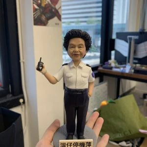 一款女性人像公仔站在灰色圓形底座上，底座前有白底黑字的牌子；公仔右手持對講機、左手自然垂放，身穿白色短袖制服襯衫（有肩章與口袋）與深色長褲、黑色皮鞋，袖臂見圓形臂章。表情親切微笑，短卷髮造型。背景為室內辦公環境，有窗外城市建築、桌面與紙箱雜物可見；一隻手掌托住底座。