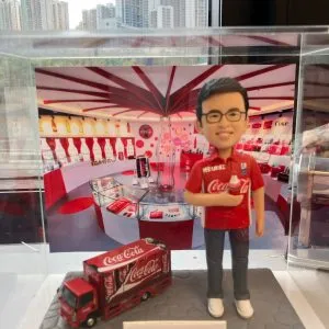 人像公仔主題場景：佩戴眼鏡的男性公仔身穿紅色可口可樂主題上衣，右手拿著可樂瓶，腳邊有縮小版Coca‑Cola貨車模型；底座前方有中文銘牌，背景為紅白相間、擺滿可樂紀念品與瓶款的展示空間，置於透明展示盒內，窗外可見高樓景致。