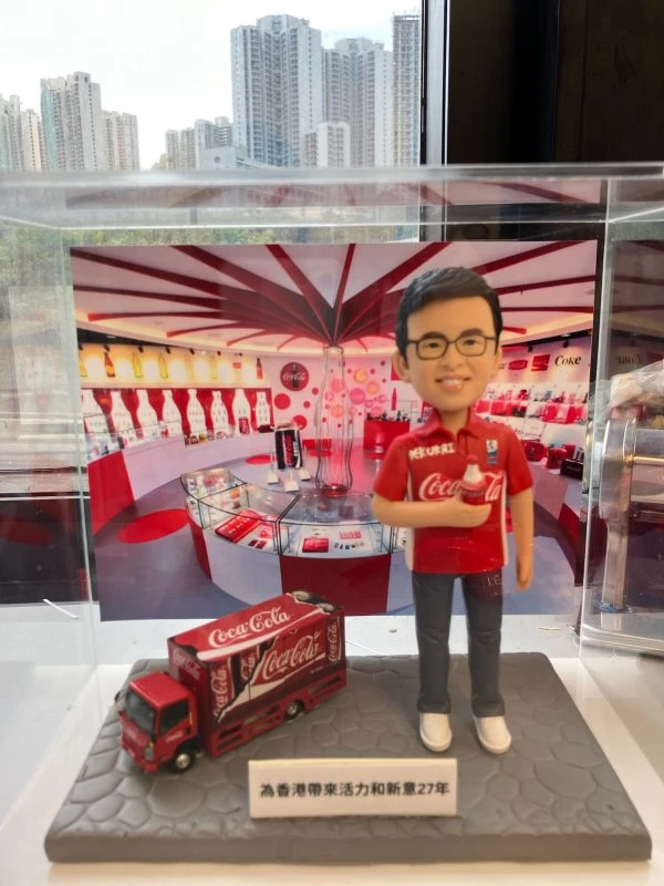 人像公仔主題場景：佩戴眼鏡的男性公仔身穿紅色可口可樂主題上衣，右手拿著可樂瓶，腳邊有縮小版Coca‑Cola貨車模型；底座前方有中文銘牌，背景為紅白相間、擺滿可樂紀念品與瓶款的展示空間，置於透明展示盒內，窗外可見高樓景致。