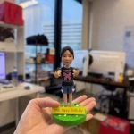 手掌心托住的小朋友主題人像公仔：bb造型女孩髮梳中分低馬尾，穿黑色上衣印有大型粉色蝴蝶結圖案與白字樣，下身藍色短褲，赤腳站在圓形草綠底座上；底座前緣附黃色牌寫著「10th Happy Birthday」；背景為室內辦公室環境，窗外大樓與桌面電腦設備散景。