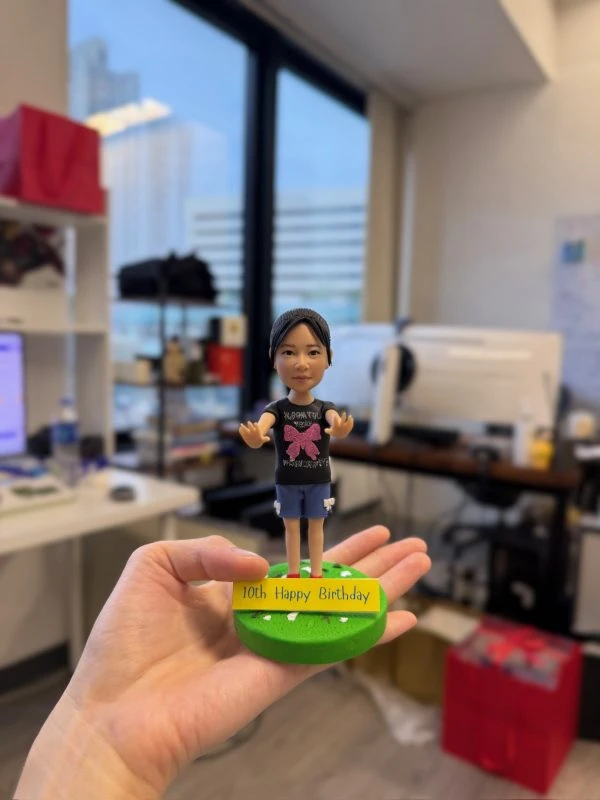 手掌心托住的小朋友主題人像公仔：bb造型女孩髮梳中分低馬尾，穿黑色上衣印有大型粉色蝴蝶結圖案與白字樣，下身藍色短褲，赤腳站在圓形草綠底座上；底座前緣附黃色牌寫著「10th Happy Birthday」；背景為室內辦公室環境，窗外大樓與桌面電腦設備散景。