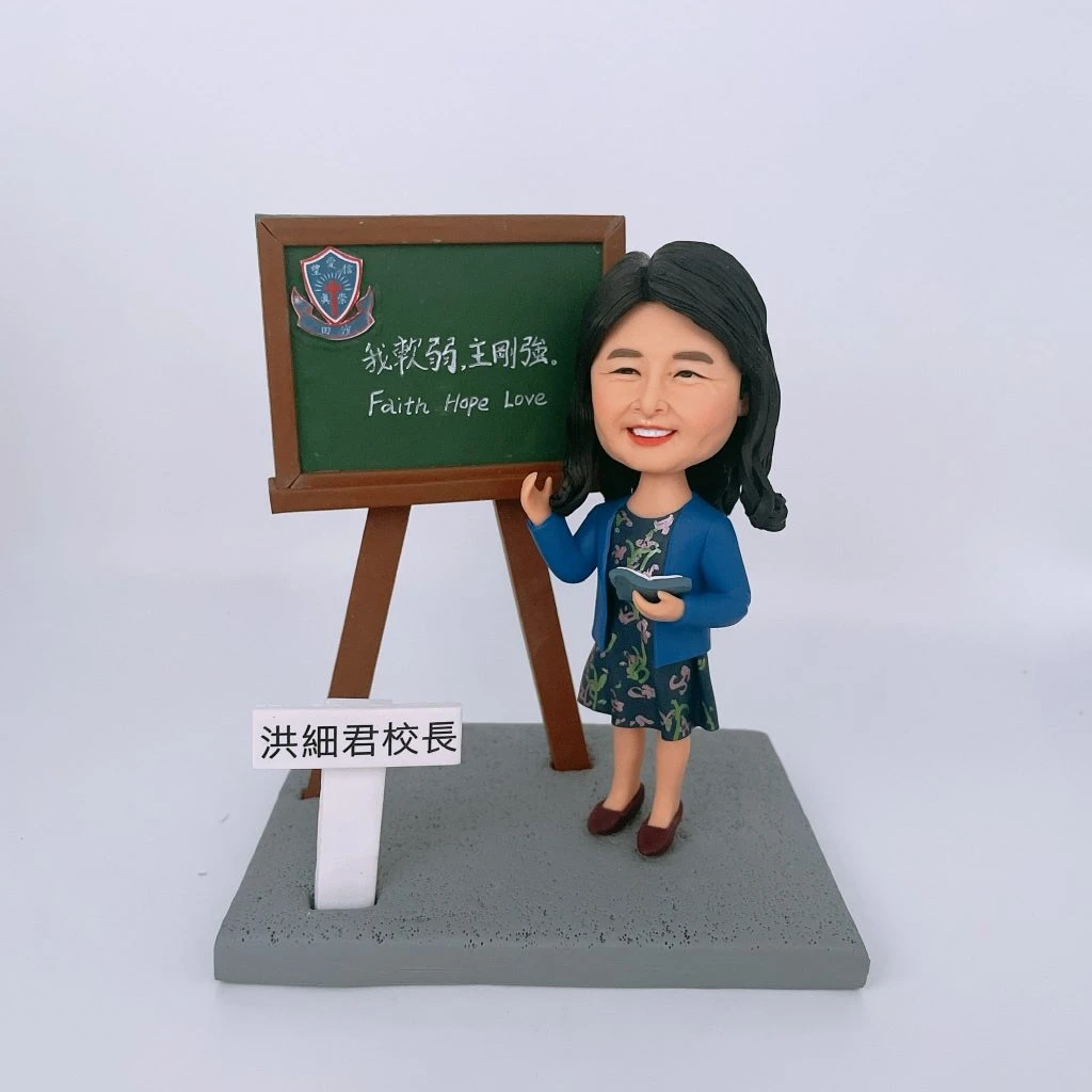 教學場景人像公仔擺設：一位女性公仔站在灰色矩形底座上，黑色長髮、面帶微笑，穿藍色外套與花卉連身裙，右手指向黑板、左手持打開的小書。旁邊立有木架黑板，上方貼有藍紅相間的校徽貼紙，黑板以中英文字寫著「我軟弱，主剛強。Faith Hope Love」。底座前方豎立白色小牌，黑字「洪細君校長」。