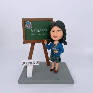 教學場景人像公仔擺設：一位女性公仔站在灰色矩形底座上，黑色長髮、面帶微笑，穿藍色外套與花卉連身裙，右手指向黑板、左手持打開的小書。旁邊立有木架黑板，上方貼有藍紅相間的校徽貼紙，黑板以中英文字寫著「我軟弱，主剛強。Faith Hope Love」。底座前方豎立白色小牌，黑字「洪細君校長」。