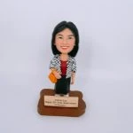 一款女性人像公仔站在木質感不規則底座上，前方金色銘牌刻有祝賀25週年與公司名稱字樣；公仔黑色短髮、微笑表情，身穿酒紅色上衣、黑色長裙與黑白格紋外套，右手挽橙黃色手袋，腳穿淺色鞋。背景純白，整體呈公司獎座風格的人像公仔。