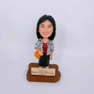 一款女性人像公仔站在木質感不規則底座上，前方金色銘牌刻有祝賀25週年與公司名稱字樣；公仔黑色短髮、微笑表情，身穿酒紅色上衣、黑色長裙與黑白格紋外套，右手挽橙黃色手袋，腳穿淺色鞋。背景純白，整體呈公司獎座風格的人像公仔。
